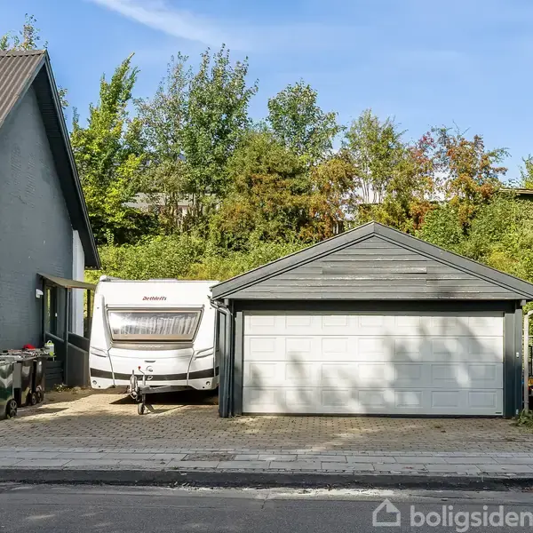 En campingvogn står parkeret ved siden af et gråt hus med garage og en sort bil. Omgivet af træer og grønt i et roligt boligområde.