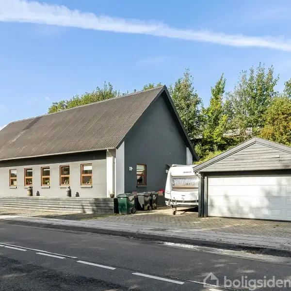 En grå bygning med skråt tag står ved siden af en hvid garage. Ved siden af bygningen ses en parkeret campingvogn og omgivelserne omfatter et vejstykke og træer i baggrunden.
