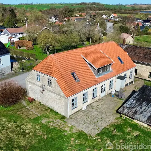 Et hus med rødt tegltag står stille blandt landlige omgivelser. Omkring det er en have og flere bygninger, mens små veje og huse er synlige i baggrunden.