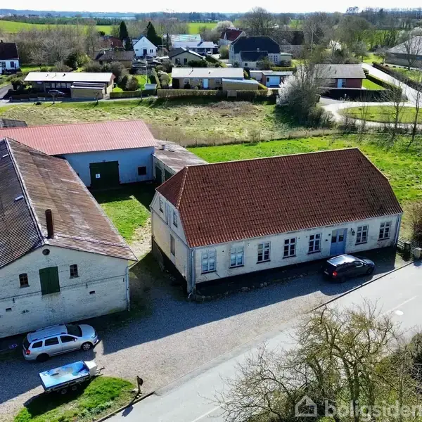 Bygning med rødt tag ved siden af en anden bygning ses fra oven. Flere små huse omkranser området i et grønt landskab med veje og biler parkeret.