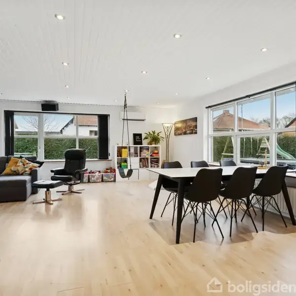 Stue med sort sofa og lænestol i venstre side, spisebord med seks stole i højre side, lyse trægulve og store vinduer med udsigt til have. Reoler med bøger langs bagvæggen.