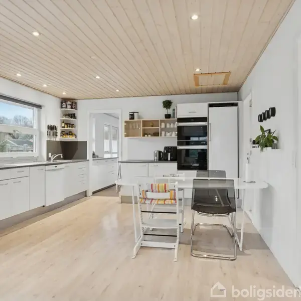 Et moderne hvidt køkken med åbne hylder, køleskab og ovn. Bordet har klare stole. Store vinduer giver udsigt til grønne omgivelser. Lyst trægulv og træloft med spotlys.