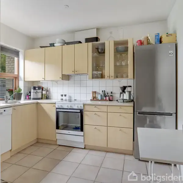 Køkken med lyse træskabe og hvid flisevæg, indeholder en ovn, opvaskemaskine, og stål køleskab. Bordpladen er dækket med små apparater, og vinduet giver naturligt lys.