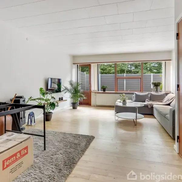 En stue med sofa og sofabord, hvor et tv er monteret på væggen. Store vinduer lader lys komme ind. Flyttekasse med teksten "jem & fix" på gulvet.