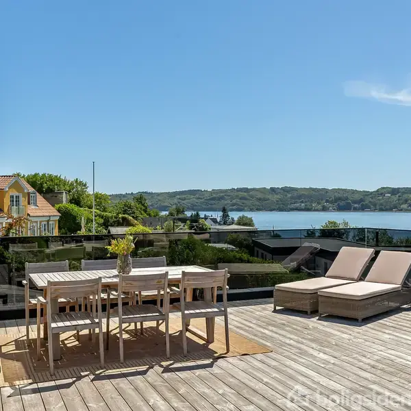 En træterrasse har havemøbler med et bord og stole samt to liggestole. I baggrunden ses en udsigt over vand og bakker under en klar, blå himmel.