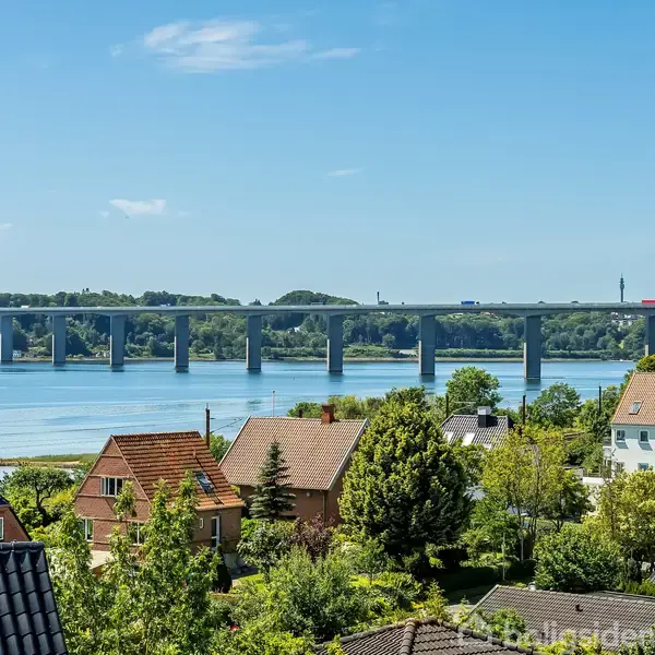 Bro strækker sig over en bred flod med biler kørende på den. Forgrunden har hustage og træer, mens baggrunden viser skovklædte områder under en klar blå himmel.