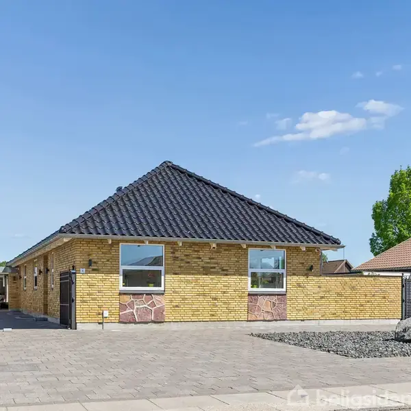 Et gult murstenshus med sort tag står på en flisebelagt gårdsplads. Til venstre ses en carport med en bil. Grønt træ i baggrunden under en blå himmel.