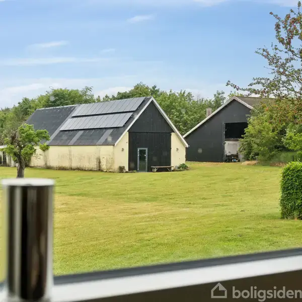 To lader med solpaneler på tage står på en stor, grøn græsplæne omgivet af træer. Set gennem et vindue med uskarpe metalgenstande i forgrunden.