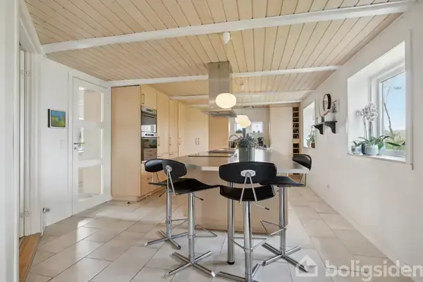 Et moderne køkken med en stor køkkenø omgivet af tre sorte barstole. Loftet er beklædt med lyst træ, og væggen har et vindue, der giver naturligt lys.