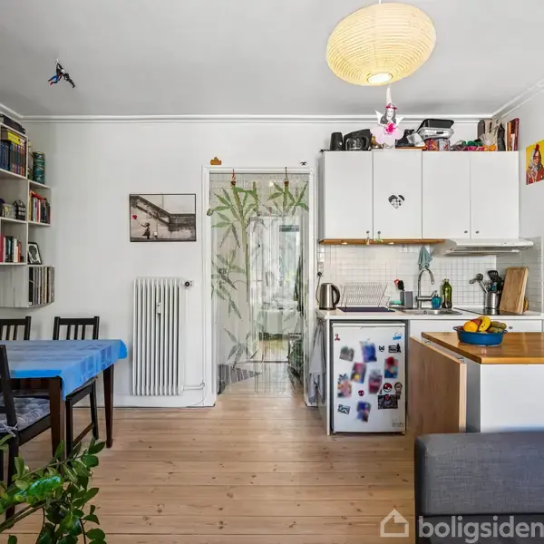 Et lille køkken med hvide skabe og en spiseplads med blå dug. Der er bøger på hyldene, planter, og et lysende loftslampe. En gennemskinnelig dør bagved viser en gang.