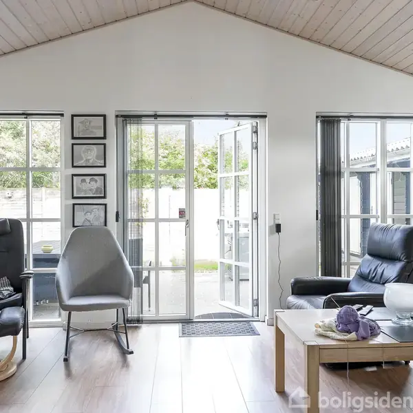 Stue med to lænestole og et bord, hvor der står en vase. Dobbelt glasdøre åbner til terrasse. Træloft og lyse trægulve omgivet af vinduer.