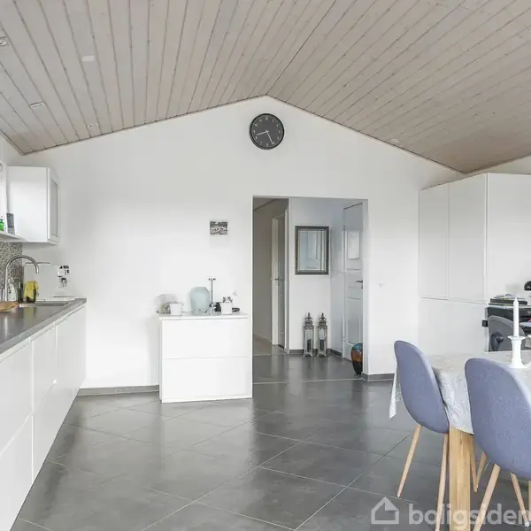 Et moderne køkken-alrum med hvide skabe og sort bordplade. Et spisebord med blå stole står til højre, omgivet af dekorationer og et ur på væggen.