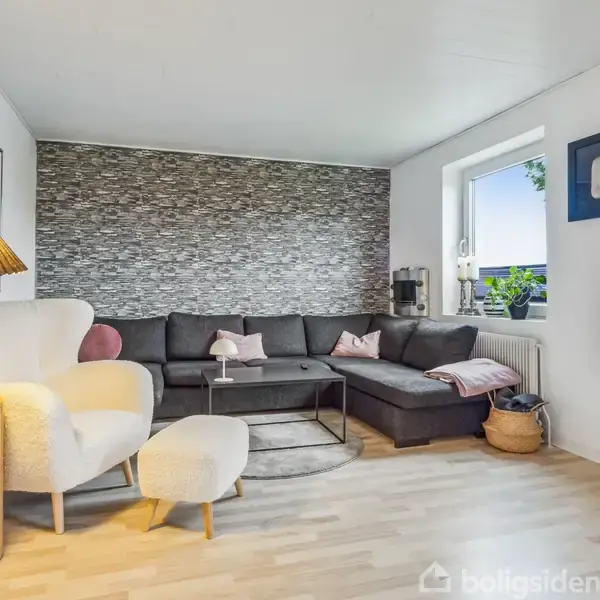 En stue med en mørkegrå hjørnesofa fyldt med puder, placeret foran en stenvæg. Til venstre står en hvid lænestol med en lampe ved siden af. En fladskærms-tv hænger på den hvidmalede væg. Et vindue med planter giver udsyn til det fri.