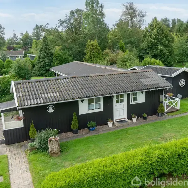 Et sort træhus med hvide vinduer og døre står stille omgivet af en velplejet have og tæt skov. Der er en parkeret bil ved siden af huset.
