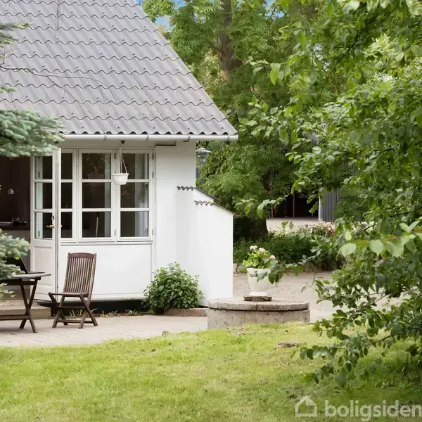 Et lille, hvidt hus står stille i en frodig have med grønne træer. Ved siden af huset står et bord og to stole på en terrasse, omgivet af buske.