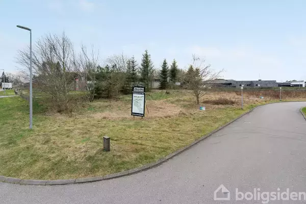 Et græsareal med et skilt i midten, omgivet af buske og træer. I baggrunden ses en række bygninger. Tekst på skiltet: "Udstillingsgrund".