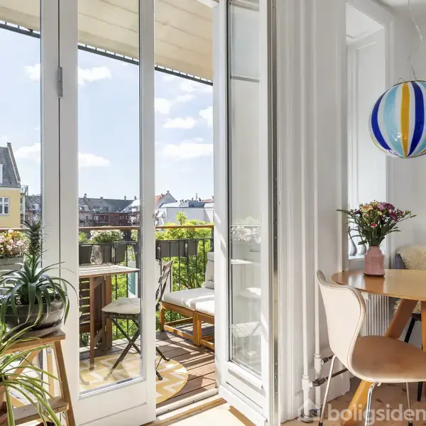 En stue med et rundt bord og stole foran åbne franske døre, der fører ud til en balkon med planter. En farverig lampe hænger fra loftet i dagslys.