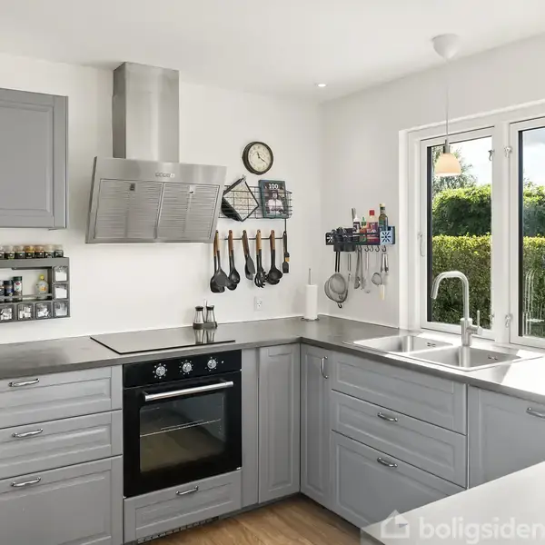 Et moderne køkken med grå skabe og en indbygget ovn. Køkkenredskaber hænger fra en stang over komfuret. Store vinduer giver udsyn til en grøn have med bil.