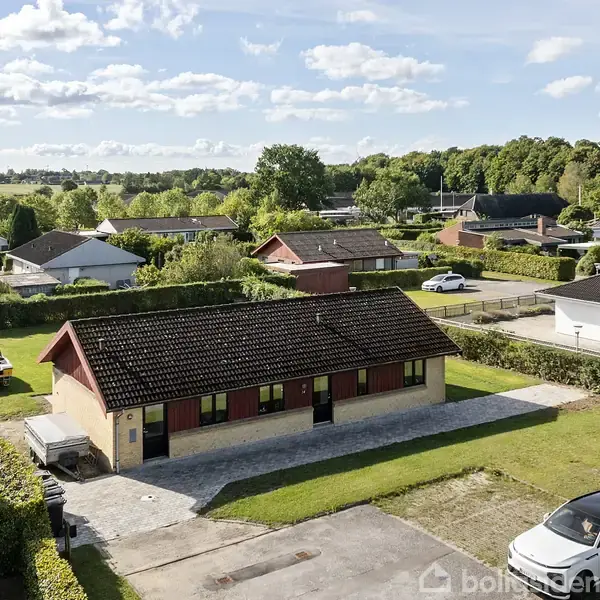 En gul murstensbygning med mørkt tag står centralt, omgivet af grønt græs og lave hække. I baggrunden ses et villakvarter med spredte huse og en grøn horisont.