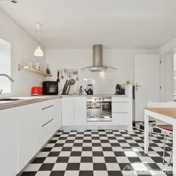 Et lyst køkken med hvide skabe står stille. Sort-hvidt ternet gulv og moderne apparater omkranser en lille spiseplads med to stole. Vinduet bringer naturligt lys ind.