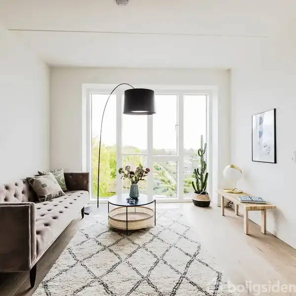 En stue med en grå sofa står ved et stort vindue. Et rundt bord med blomster foran sofaen, en gulvlampe bøjer sig over bordet. En kaktus står i hjørnet.