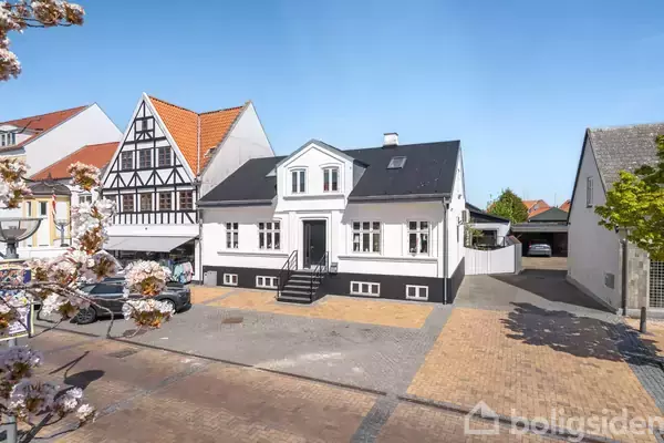 Hus står roligt i en bymæssig kontekst omgivet af andre bygninger. Det er hvidt med sort tag og detaljer, har en trappe foran og flisebelagt gårdsplads. Blomstrende træer og blå himmel omgiver det.