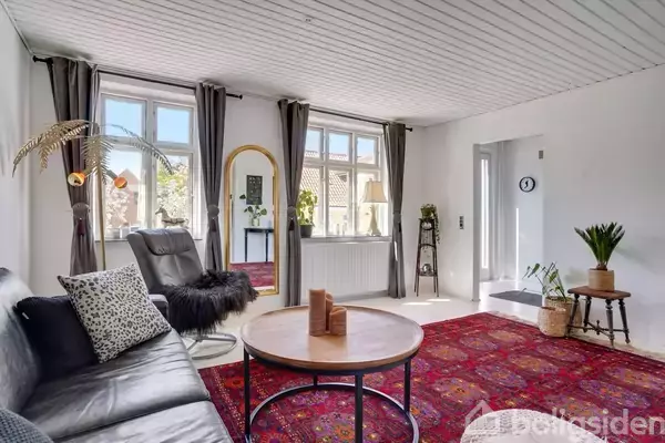 En lys stue med sort lædersofa, rundt sofabord og rødt orientalsk tæppe. To store vinduer med gardiner lader lys ind. Spejl og planter pryder rummet. Ingen tekst.