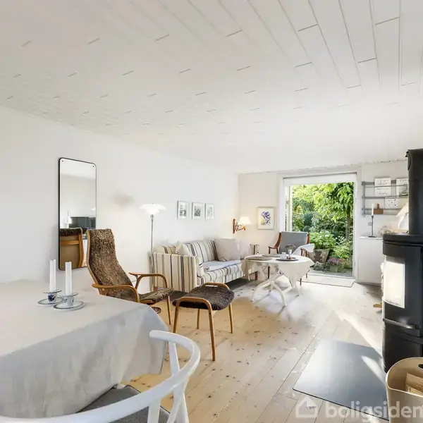 En stue med lyse trægulve indeholder en pejs, sofaer og lænestole. Rummets bagvæg har et stort vindue med udsigt til en frodig have. Et spisebord står til venstre.