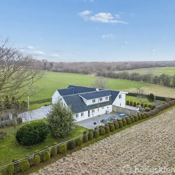 Et hvidt hus med blå tag sidder stille i et åbent landskab omgivet af grønne marker og træer under en lyseblå himmel med få skyer.