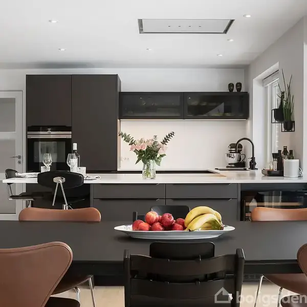 Køkkenbord med frugtskål fyldt med bananer og æbler. Bagved ses et moderne køkken med sorte skabe, en vase med blomster, og stearinlys. Vindue til højre slipper lys ind.