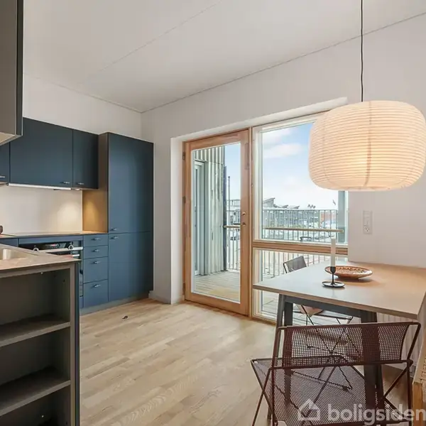 Et moderne køkken med blå skabe og hvide bordplader. Ved siden af er et spiseområde med et bord, to stole og en stor lampe. En glasdør fører ud til en altan.
