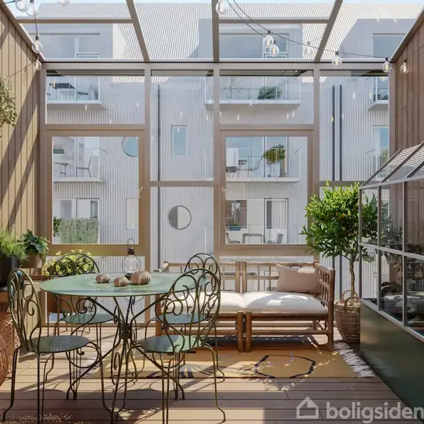 Et glasoverdækket terrasseområde med et rundt metalbord og fire stole. En sofabænk står ved vinduerne, omgivet af planter. Bygninger ses udenfor gennem ruderne.