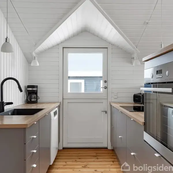 Et moderne køkken med grå skabe langs to vægge og en trædør i enden. Bordpladerne har træfinish. Der er indbyggede ovne og en kaffemaskine. Væggene er hvidmalede træpaneler.
