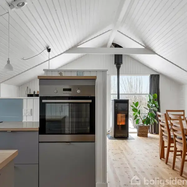 Køkkenet har en indbygget ovn og køkkenø. I stuen brænder en brændeovn, omgivet af spisebord og stole. Rummets hvide vægge og skrå loft skaber lys og åbenhed.