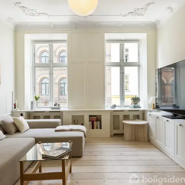 Stue med grå sofa og glasbord ved siden af en stor fladskærms-tv på et hvidt skab. Store vinduer giver naturligt lys, med udsigt til en murstensbygning.
