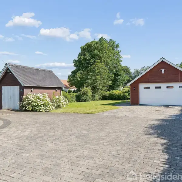 En stor flisebelagt indkørsel foran to garager; den ene rød med hvide porte, den anden brun med buske ved siden af, omkranset af grønne træer og en velholdt have.