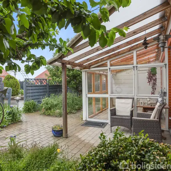 En overdækket terrasse med siddemøbler og glasvægge, omgivet af en frodig have med stier og grønne planter. En cykel og et plankeværk ses i baggrunden.