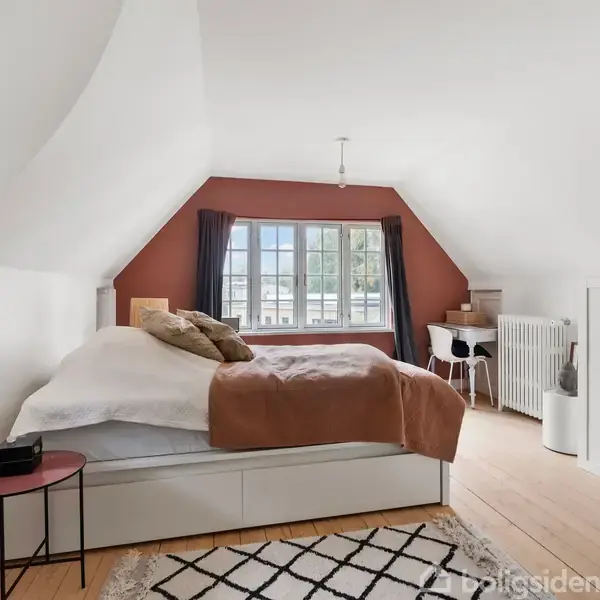 Et soveværelse med en stor seng dækket af puder og et tæppe, står ved siden af et lille bord. Forrest ses et skrivebord ved et vindue med røde gardiner.