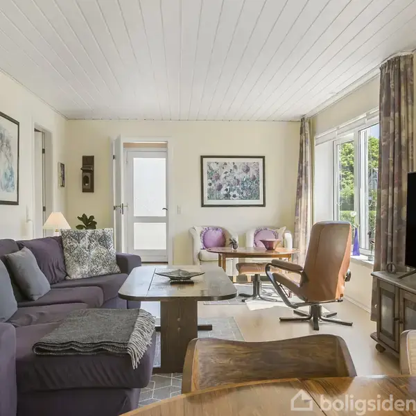 En stue med en lilla sofa fyldt med puder står ved siden af et træbord. En stor fladskærms-tv står på et bord foran. Lyse vægge med billeder fuldender rummet.