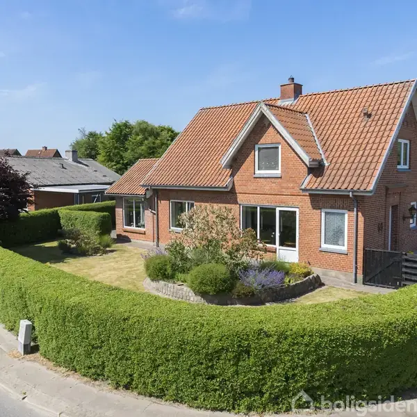 Rødt murstenshus med rødt tegltag omgivet af en velplejet hæk og have. Huset står på hjørnet af en vej med et vejskilt der viser "Munkevænget". Klar, solrig himmel.