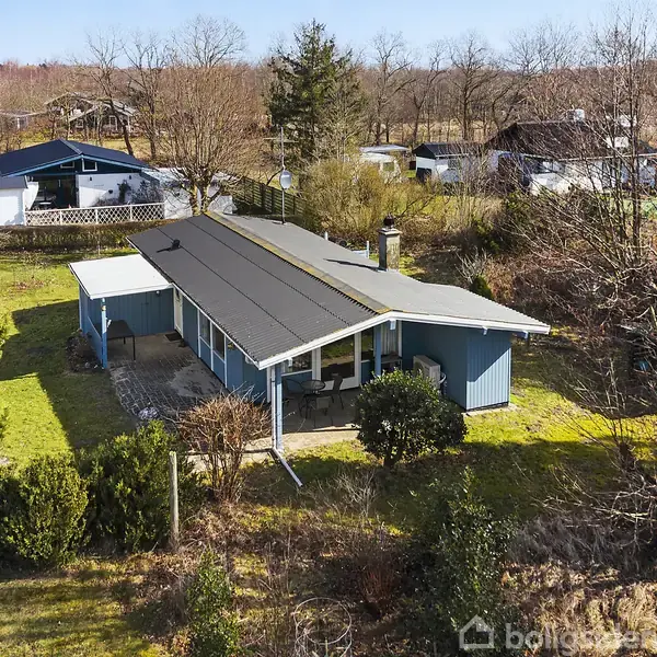 Et blåmalet sommerhus med sort tag står omgivet af træer og buske. Huset har en veranda og græsplæne i forhaven. Det ligger i et bebygget skovområde med andre huse.