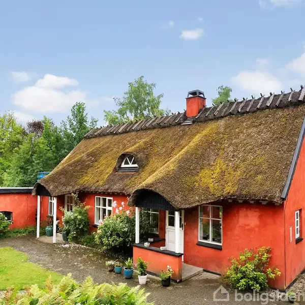 Rødt, stråtækt hus med flere vinduer, omgivet af grønne buske og træer. Grussti fører til hoveddøren, og der er plantekrukker langs facaden. Klar blå himmel ses i baggrunden.
