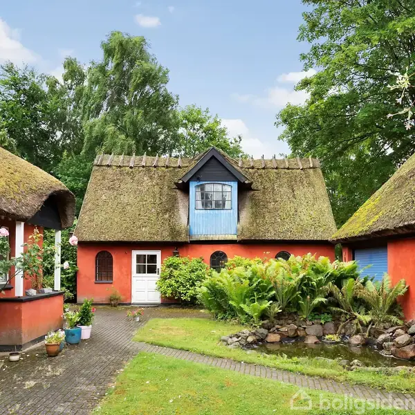 Et charmerende, stråtækt hus står i en have med grønne planter og blomster. Stedet er omgivet af gamle træer og har en blå dør og vindue i øverste etage.