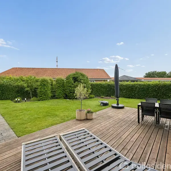 Træterrasse med sorte stole og parasol, står ved siden af en grøn græsplæne og fliser. Omkranses af tætklippet hæk og rødt tegltag ses i baggrunden under en blå himmel.