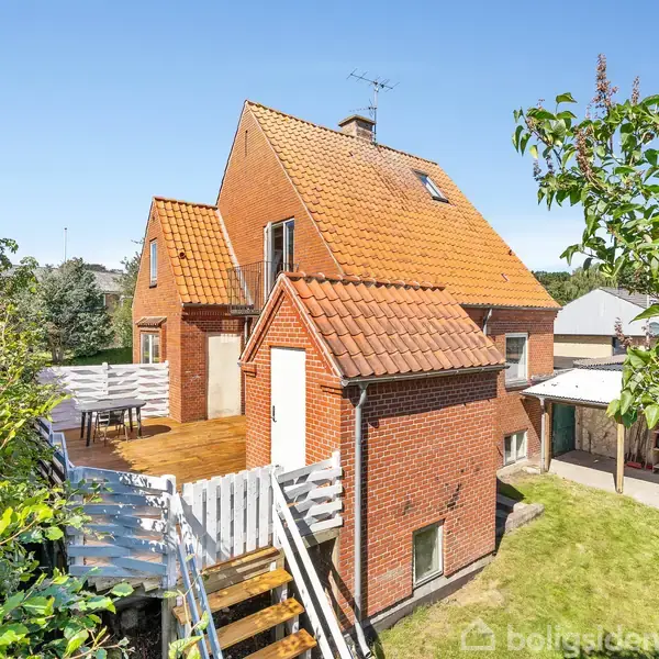 Et rødt murstenshus med rødt tegltag har en træterrasse med hvide møbler. Huset er omgivet af en have med træer og buske under en blå himmel.