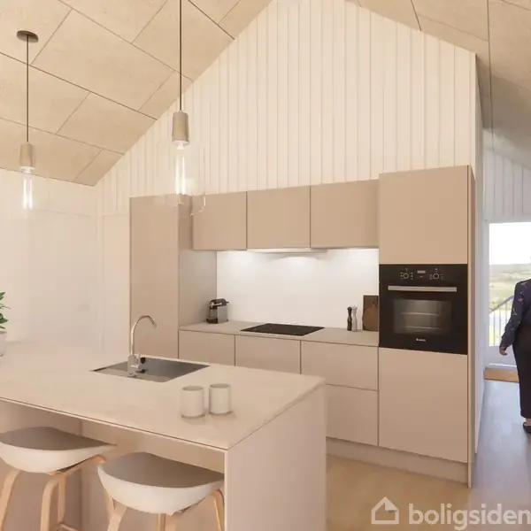 Et moderne køkken med lyse skabslåger og en central ø med vask og to barstole. En person står i en gang ved siden af, mens store vinduer bringer lys ind.