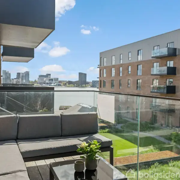 Grå sofa står på altan med glasrækværk og udsigt til moderne lejlighedsbygninger. Kaktusser og en grøn plante pryder et bord. Bylandskab er synligt i baggrunden.