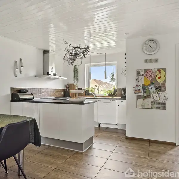 Et køkken med centralt placeret hvid køkkenø under en udsmykning fra loftet. Spisestue til venstre, udsigt gennem stor vindue, vægdekorationer og original kunst på højre væg.