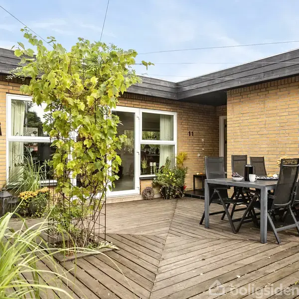 Terrassen har et bord med seks stole arrangeret til en udendørs middag. En murstensbygning danner baggrunden, omgivet af grønne planter og en klar himmel. Nummer 11 er på væggen.