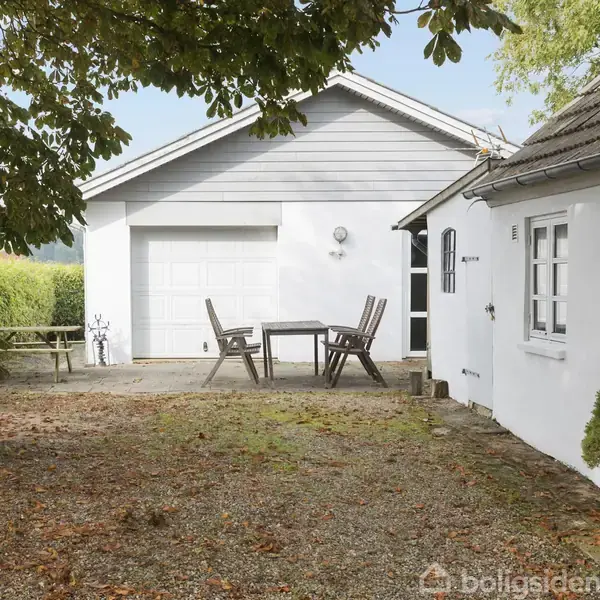 En hvid bygning med gråt tag og en gårdsplads, hvor der står et bord med stole. Ved siden af ses en garage med hvid port. Omkranset af træer og hæk.